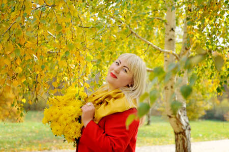 Blonde woman with yellow flowers in autumn outdoors. She is standing near a birch tree with yellow leaves in a red coat, looking at the camera and smiling. Fashion portrait. Close-up.の写真素材