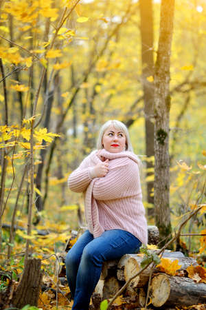A bright middle-aged woman in a delicate knitted jumper poses on a Sunny autumn day in the forest on logs. Arms crossed over his chest. He looks up dreamily. Woman's size plus xxl. Vertical view.の写真素材