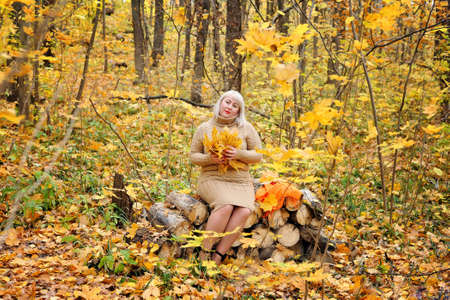 Blonde on the background of bright autumn leaves posing in a dress in a Sunny forest, smiling thoughtfully looking at the frame. Women's plus size xxl. Horizontal view. General plan.の写真素材