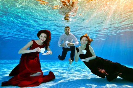 Two unusual cute girls and a guy pose and play underwater at the bottom of the pool with white cups in their hands, in dresses and a shirt on a blue background. Tea party underwater. Conceptの写真素材