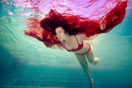 Surreal portrait of a girl who swims under the water like a bird, arms outstretched, in a red swimsuit and holding a red cloth in her hands against a bright light. Vintage processing.の写真素材