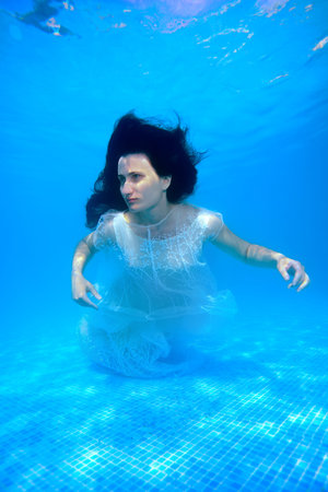 A beautiful girl in a white dress swims and poses underwater in the pool with her hair down on a...
