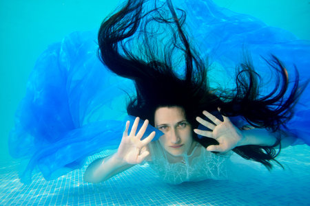 Portrait of an unusual bride who poses for the camera underwater at the bottom of the pool with her long hair down, in a white dress with a blue cloth in her hands. Portrait. Surrealism.の写真素材