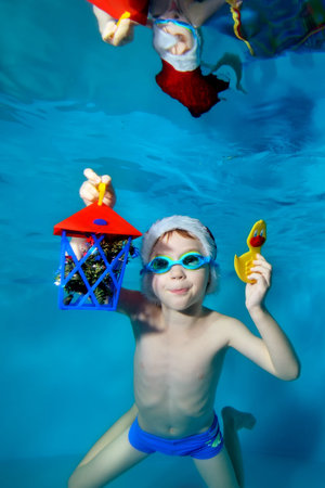A little boy poses underwater with Christmas toys in his hands, wearing a red Santa hat and swimming goggles. He looks at the camera and smiles at the bottom of the pool. Christmas. Portrait. Concept.の写真素材