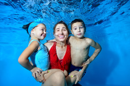 A happy family: an adult mother, her little son and little daughter play under the water in the pool, they smile and pose for the camera on a blue background surrounded by bubbles. Closeup.の写真素材