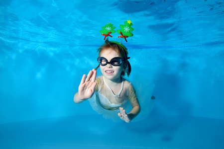 A little girl with Christmas decorations poses for the camera underwater in a pool on a blue background. The Concept Of Christmas. Portrait. Horizontal viewの写真素材