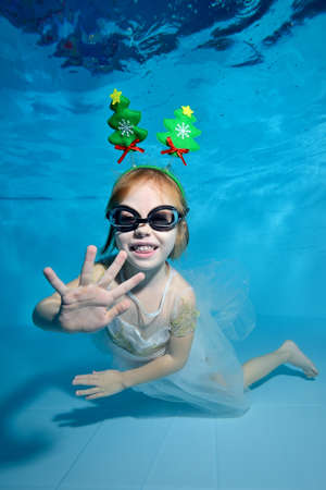 Portrait of a little girl with Christmas decorations in the form of a Christmas tree on her head. She poses for the camera under the water in the pool on a blue background and smiles. Vertical viewの写真素材