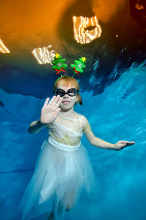 A little girl with Christmas decorations in the form of a Christmas tree on her head poses for the camera underwater in a childrens pool on a blue background. The Concept Of Christmas. Portraitの写真素材