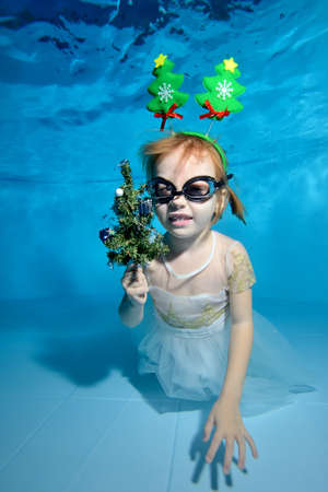 A little girl with a small toy Christmas tree in her hand and a white dress poses for the camera underwater in a pool on a blue background and smiles. Portrait. Vertical viewの写真素材