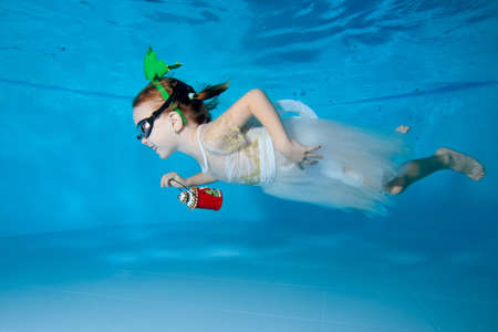 Portrait of a beautiful little girl with a Christmas toy in her hand and a white dress. She swims underwater in a pool on a blue background and smiles. Horizontal orientationの写真素材