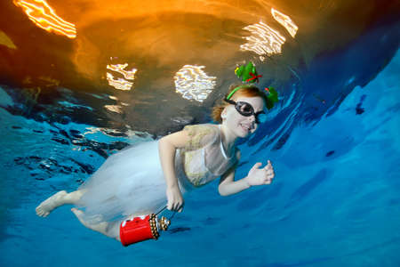 A child, a girl, with a red flashlight in her hand, smiles and swims under the water in a childrens pool in a white dress and swimming glasses. Portrait. Horizontal orientation of the imageの写真素材