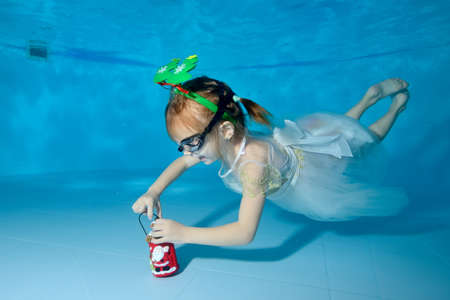 A happy little girl in a white dress dives for a Christmas toy at the bottom of the childrens pool and smiles. Portrait. Horizontal orientationの写真素材