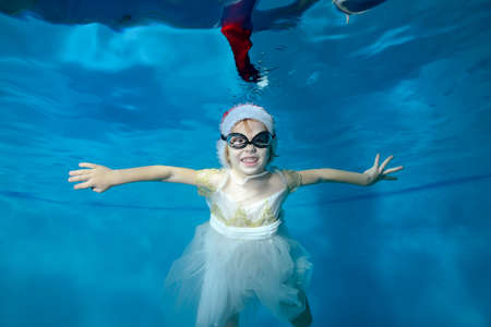 Portrait of a cheerful little girl in a Santa hat and white dress, who swims and poses for the camera underwater in a childrens pool and smiles. Concept. Horizontal orientationの写真素材