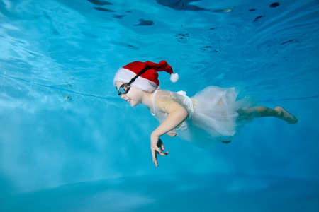 A child, a little girl, in a white dress and a Santa Claus hat, swims under the water in the pool and smiles. Portrait. Side view. The Concept Of Christmas. Horizontal orientationの写真素材