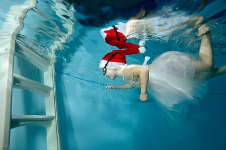 A cute little girl, wearing a Santa hat and a white dress, swims under the water to the white stairs in the childrens pool and smiles. Portrait. Side view. Horizontal orientationの写真素材