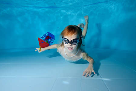 A little girl with a new years gift in her hand lies under the water at the bottom of the pool in a white dress and swimming glasses. Portrait. Horizontal orientation of the imageの写真素材