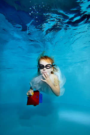 Funny child, a girl, swims and poses with a childs gift under the water in the pool in a white dress on a blue background. She looks at the camera and smiles sweetly. Portrait. Vertical viewの写真素材