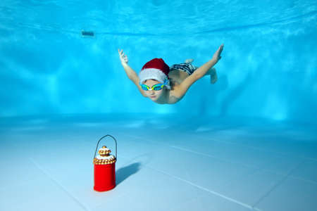 A little boy in a red Santa hat dives to the bottom of the pool for a Christmas toy. Concept. Portrait. Horizontal viewの写真素材