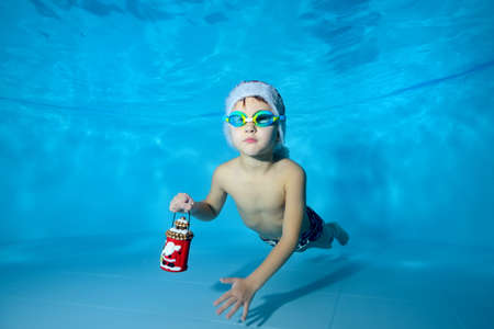 A cute little boy poses for the camera in a red Santa hat and a Christmas toy in his hand underwater at the bottom of a childrens pool. Concept. Portrait. Horizontal orientationの写真素材