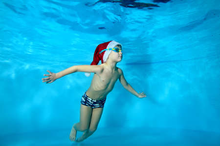 A charming child, a boy, swims under the water in the pool in a red Santa hat with his arms outstretched. Portrait. Concept. Horizontal orientation of the photoの写真素材