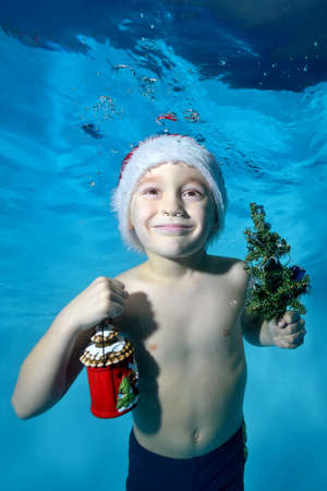 Portrait of a little boy in a red Santa Claus hat with a new year's gift in his hand and a small Christmas tree, who poses under water on a blue background. Vertical orientation of the photo.の写真素材
