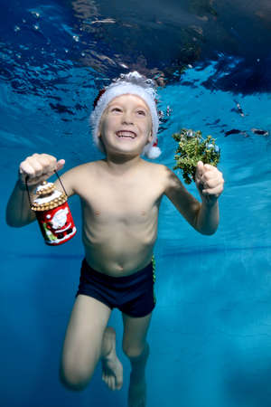 Laughing little boy swims and plays underwater in the pool in a red Santa hat with Christmas gifts in hand on a blue background. Portrait. Vertical orientation of the photo.の写真素材