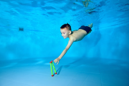 Cute athletic boy dives to the bottom of the pool for a toy. Fun dives underwater. Active happy child. Swimming classes. Bodily exercises. Healthy lifestyleの写真素材