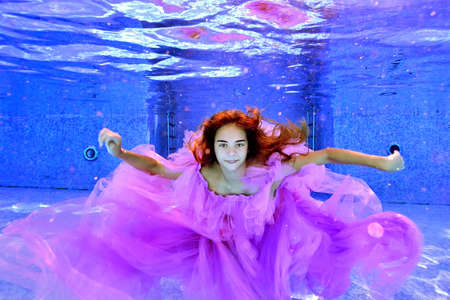 Portrait of a teenage girl in a beautiful dress, who poses underwater in an outdoor pool with her arms outstretched. Her long hair flutters in the water. Horizontal orientation.の写真素材
