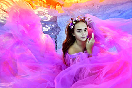 Portrait of a cute teenage girl posing underwater at the bottom of an outdoor pool in a beautiful dress and a wreath of flowers. Underwater photography. Horizontal orientation.の写真素材