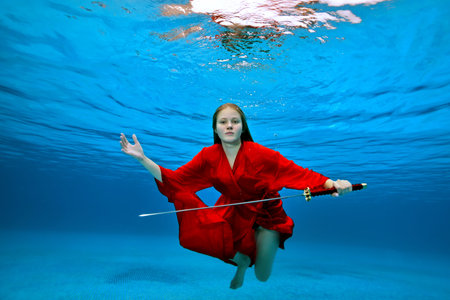 A beautiful girl in a red kimono poses for the camera underwater in a pool with a samurai sword on a...