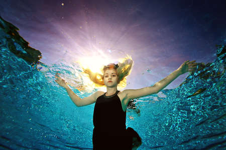 A surreal portrait of a young blonde girl swimming underwater in a pool in a black dress, arms outstretched like a bird. Shooting from the bottom of the pool in the contour light of the sun.の写真素材