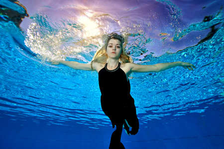 Beautiful blonde girl swims under the water in the pool in a black dress. She floats in the water, arms outstretched, like a bird against the setting sun. Bottom view. Underwater photography.の写真素材