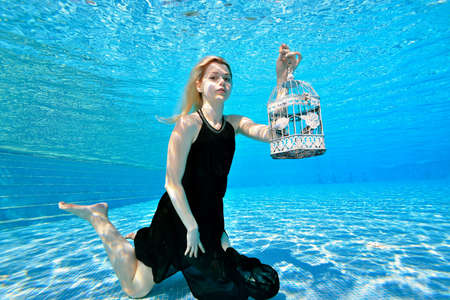 Fashionable portrait of a blonde girl underwater. She poses in an outdoor pool in a black dress and holds a white check. Bright colors. Underwater photography.の写真素材