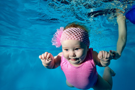 Portrait of a charming, breastfed baby who swims underwater in a childrens pool in a pink swimsuit and with a bow on her head. Mom-coach insures her in the water. Healthy lifestyle. Concept.の写真素材