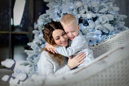 A cute little boy hugs his mom tightly while sitting on a couch against the background of a beautiful Christmas tree decorated with toys and garlands. Happy New Year concept. Horizontal view.の写真素材