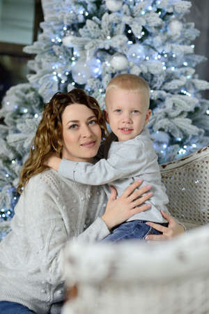 A beautiful young mother and her little son, hugging each other, look into the camera and smile, sitting against the background of a Christmas tree decorated with toys and garlands. Concept. Portrait.の写真素材