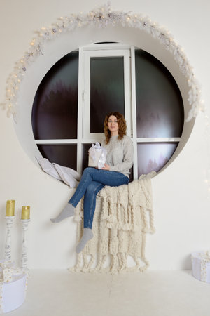 A beautiful young woman with a gift box in her hands is sitting at home on a round window decorated with garlands and yellow lights, looking at the camera and smiling. Portrait.の写真素材