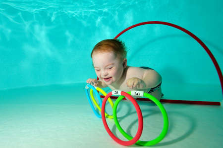 A happy kid with Down syndrome swims and collects toys underwater from the bottom of a childrens pool during a sports activity. Childrens disability. Concept. Portrait. Horizontal orientation.の写真素材