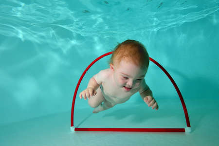 A cute little boy with Down syndrome plays in the water and swims through a hoop at the bottom of the pool. The concept of a happy childhood of a child with a disability. Horizontal orientation.の写真素材