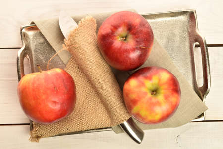 Three red juicy fresh apple with a knife and a jute napkin on a metal tray on a painted wooden table.の写真素材