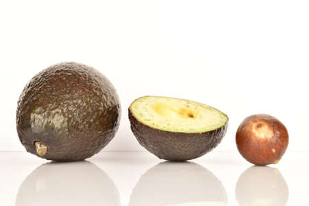 One whole and one half, dark green, ripe fragrant tasty avocados, next to it there is one whole brown stone from the avocado. The background is white.の写真素材