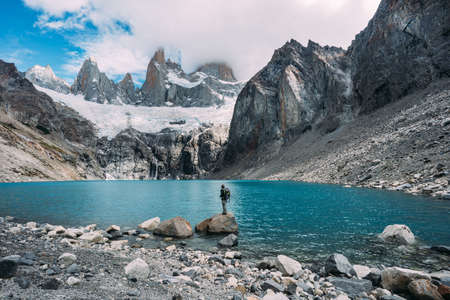 One small man backpacker standing in front of a blue lake and mountains covered with snowの写真素材