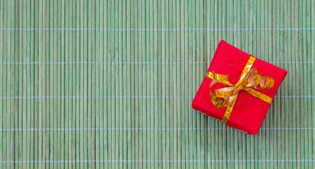 Gift box packed in red wrapping paper and decorated with golden ribbon placed on green bamboo matの写真素材