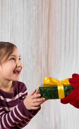 Excited smiling little girl is getting decorated gift box of hands of Santa Claus dressed in...