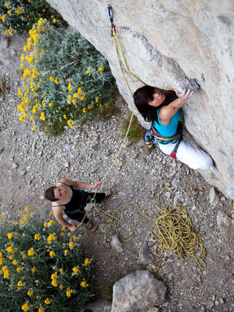 Team of climbers. Two female climbers work as a team. One leads climbs the wall another belays her keeping the rope. Nice yellow flowers on the backgroundの写真素材