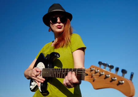 Stylish female musician with red hairs wearer in black hat and green shirt plays on the guitar. Deep blue sky on the background. Focus on the faceの写真素材