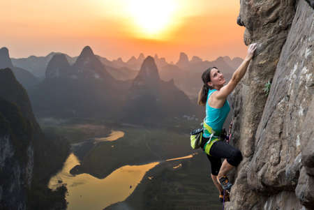 Female extreme climber conquers steep rock against the sunset over the river. China typical Chinese landscape with mountains and river