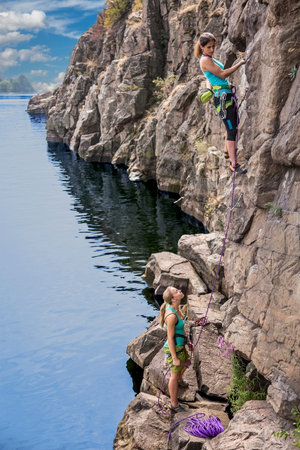 Two young female extreme climbers climb the rock. One is leading another is belaying. Rocky beach river natural rock outdoor summerの写真素材