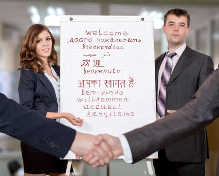 Two young corporate trainers welcome participants of the training They use flip chart with WELCOME sign written in many different languages Handshaking people on the foreground.の写真素材