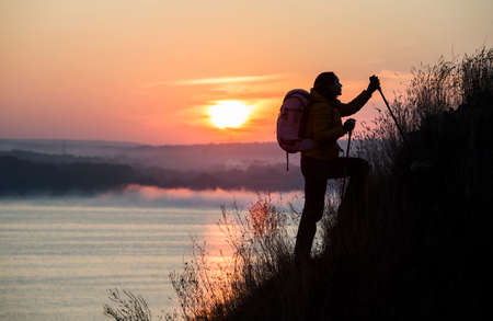 Backpacker is climbing the steep slope Sunrise and river on the backgroundの写真素材